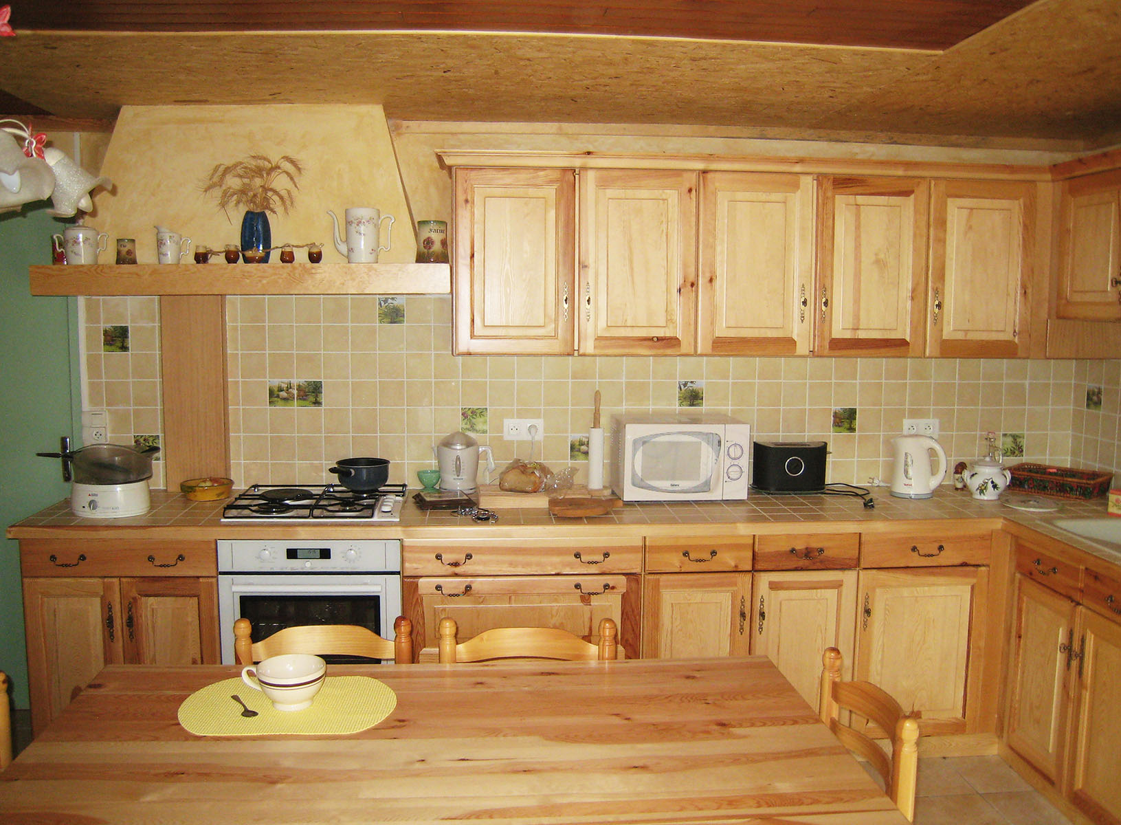 Façade — Pin vernis.
Poignées — Serrures et poignées tombantes.
Plan de travail — Carrelage avec alaise bois.
Électroménager — Four, plaque de cuisson, hotte.